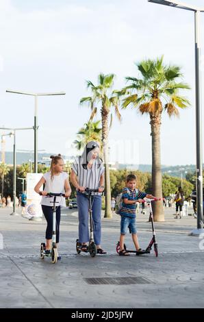 Fröhlicher Familienleben und Urlaubskonzept. Mutter, kleiner Junge, Mädchen, Roller reiten, in der Altstadt spazieren gehen, Straße. Lachen an einem sonnigen Sommertag. Stockfoto