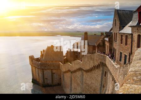Alte Festungsmauern, Häuser und Gebäude der alten Stadt-Festung von Mont Saint-Michel in den Strahlen der untergehenden Sonne gegen das Ufer des Stockfoto