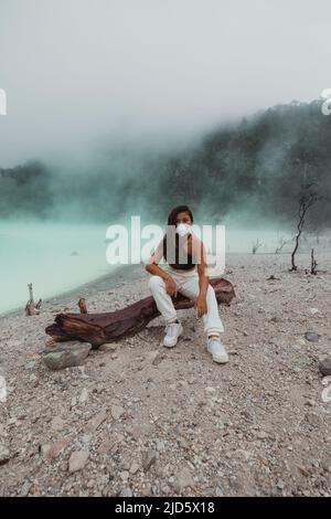 Junge Indonesierin, die am Schwefelsee in Kawah Putih sitzt und von grünem Nebel umgeben ist Stockfoto