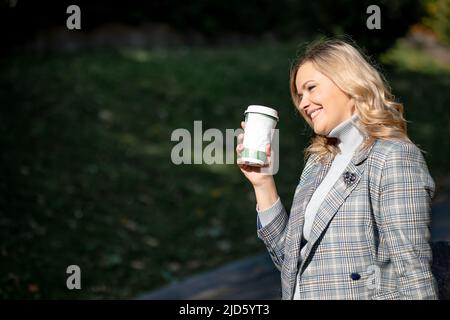 Charmante Blondine mit langen lockigen Haaren in grau-blauer Tweed-Jacke und passendem Kaschmirpullover, die eine Tasse Tee in den Händen hält, während sie bei Sonnenschein geht Stockfoto