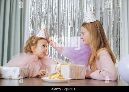 Geburtstag ist fünf Jahre alt. Die ältere Schwester passt den Karnevalshut der charmanten jüngeren Schwester, die das Festhorn bläst, vorsichtig an. Mädchen sind wunderschön Stockfoto