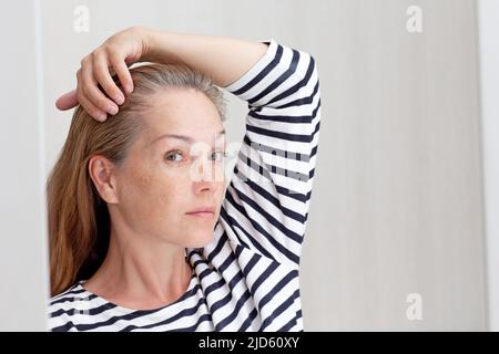 Schöne 40s Frau Blick auf graue Haare in Spiegelreflexion auf wachsende Wurzel Stockfoto