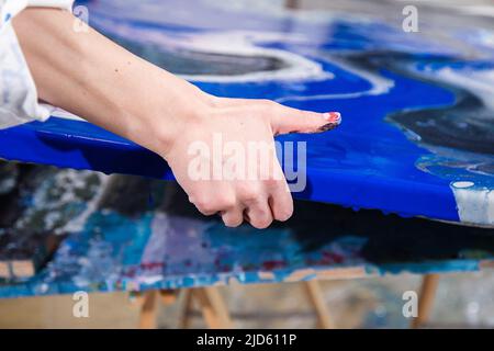 Beschnittenes Foto von Händen einer Frau, die Leinwand mit abstraktem Bild in verschiedenen blauen, weißen, schwarzen Farben auf dem Schreibtisch halten. Stockfoto