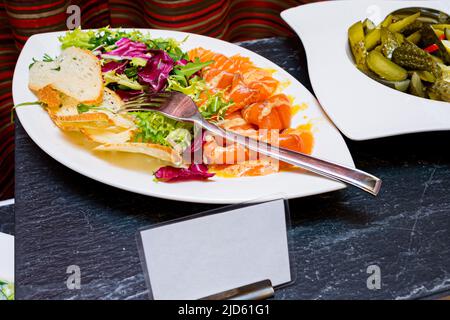 In Meersalz marinierter Lachs, Salatblätter und dünne Croutonscheiben. Ein Teller mit eingelegten Gurken in Scheiben auf einem anderen Teller. Der Buffettisch. Stockfoto