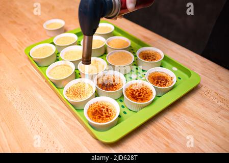 Ein funktionierendes Gasbrenner macht die Kruste von crème brûlée (creme brulee) Desserts. Ein grünes Kunststofftablett mit Portionen von crème brûlée in Ramekins auf dem Stockfoto