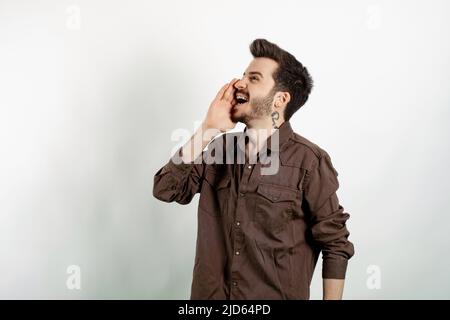 Kaukasischer junger Mann mit Hemd, der isoliert auf weißem Hintergrund posiert und laut auf der Seite mit der Hand am Mund schreit. Kommunikationskonzept. Stockfoto