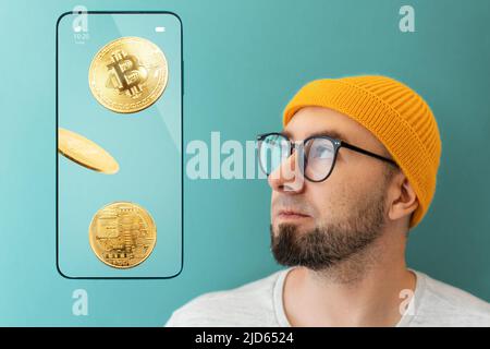 Porträt eines kaukasischen jungen Mannes mit Brille und Hut, der auf ein Smartphone schaut, das Bitcoins zeigt. Das Konzept von Banking, Kryptowährung und Finanzen. Stockfoto
