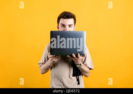 Hübscher junger Mann in legerer Kleidung, der isoliert auf gelbem Hintergrund posiert und hinter dem Laptop aus der Kamera blickt. Stockfoto