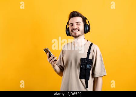 Fröhlicher junger Mann mit beigefarbenem T-Shirt, der isoliert auf gelbem Hintergrund über kabellose Kopfhörer posiert und Musik vom Smartphone hört. Stockfoto