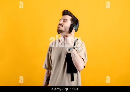 Junger Mann in beigem T-Shirt, der isoliert auf gelbem Hintergrund über kabellose Kopfhörer posiert und Audioguide meditiert. Berühren des Kopfhörers. Stockfoto