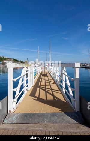 Brixham Harbour, Devon, England, Großbritannien – eine Gangway mit weißen Schienen, die vor einem tiefblauen Himmel und blauem Meer liegt. Weitwinkelaufnahme Stockfoto