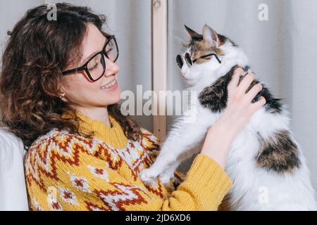 Porträt einer jungen Frau in einer Brille mit lockigen Haaren, die eine weiße Katze in einer kleinen Brille hält, die zu Hause am Bett auf dem Boden sitzt. Stockfoto