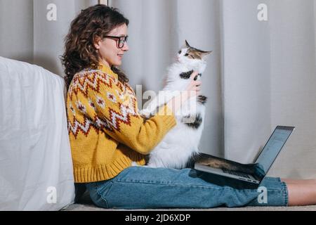 Nettes freiberufliches Mädchen in einer Brille und einem gelben Pullover arbeitet zu Hause auf einem Laptop, der auf dem Boden sitzt und ihre weiße Katze umarmt Stockfoto