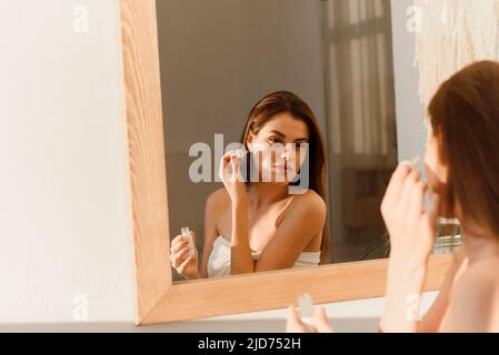 Gesichtspflege. Attraktive Frau mit Hyaluron-Gesichtsserum, feuchtigkeitsspendende und Hautpflege, stehend in der Nähe des Spiegels im modernen Innenbadezimmer. Täglich Stockfoto