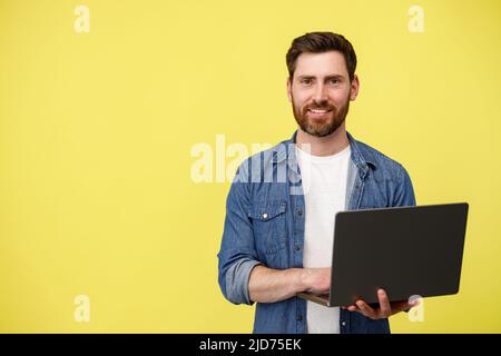 Schöner lächelnder Mann mit Bart, der Laptop hält und auf gelben Hintergrund auf die Kamera schaut Stockfoto