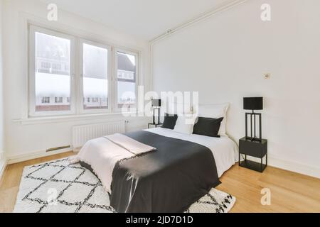 Komfortables Bett an einer weißen Wand in einem Schlafzimmer in einem modernen, minimalistischen Stil mit Fenstern Stockfoto