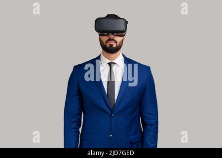 Bärtiger Mann, der im vr-Headset steht, einen Virtual-Reality-Film mit ruhigem Gesichtsausdruck, innovativer Technologie und einem offiziellen Anzug ansieht. Innenaufnahme des Studios isoliert auf grauem Hintergrund. Stockfoto