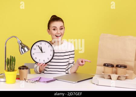 Glücklich optimistisch Frau Büroangestellte mit großer Wanduhr zeigen Finger auf Papiertüte, Kaffeetassen und Pizza, zufrieden mit der schnellen Lieferung. Studio-Innenaufnahme isoliert auf gelbem Hintergrund. Stockfoto