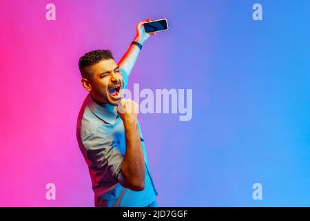 Porträt eines ausgestreunten, staunenden Mannes im Hemd, der mit einem Smartphone in den Händen steht, Livestream sendet und fröhlich schreit. Innenaufnahmen im Studio, isoliert auf farbigem Neonlicht-Hintergrund. Stockfoto