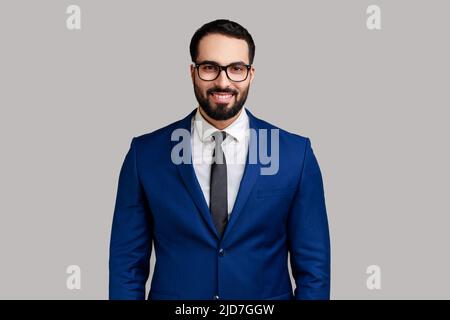 Porträt eines glücklichen lächelnden, zufriedenen bärtigen Geschäftsmannes in einer Brille, der mit positivem Ausdruck die Kamera anschaut und einen offiziellen Anzug trägt. Innenaufnahme des Studios isoliert auf grauem Hintergrund. Stockfoto