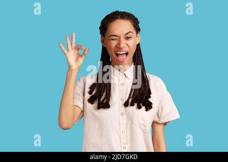 Portrait einer fröhlichen jungen erwachsenen Frau mit schwarzen Dreadlocks, die ein gutes Zeichen zeigt, versichert, dass alles in Ordnung ist, sieht gerne aus, trägt weißes Hemd. Innenaufnahme des Studios isoliert auf blauem Hintergrund. Stockfoto