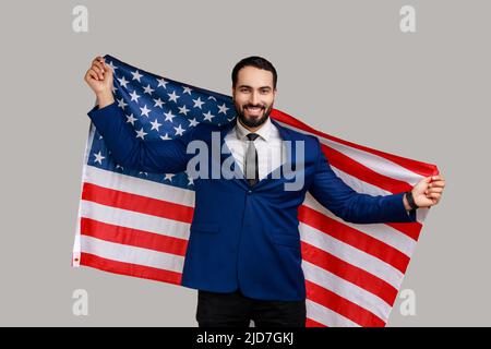 Lächelnder bärtiger Mann, der die USA-Flagge hält und fröhlich auf die Kamera schaut, den Nationalfeiertag feiert und einen offiziellen Anzug trägt. Innenaufnahme des Studios isoliert auf grauem Hintergrund. Stockfoto