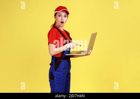 Porträt einer schockierten, erstaunten Arbeiterin, die online am Laptop arbeitet, mit offenem Mund auf das Notebook-Display zeigt und Overalls trägt. Innenaufnahme des Studios isoliert auf gelbem Hintergrund. Stockfoto