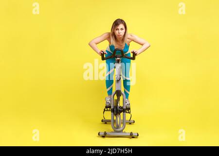 Porträt einer sportlich attraktiven Frau, die HIIT-Cardio-Training macht, Fahrrad-Intervalltraining trainiert und blaue Sportkleidung trägt. Innenaufnahme des Studios isoliert auf gelbem Hintergrund. Stockfoto