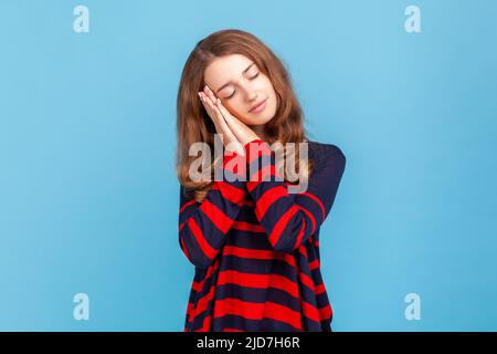 Frau in gestreiftem Pullover im lässigen Stil, schläft auf ihren Handflächen und lächelt zufrieden, hat ein bequemes Nickerchen und ruht sich aus, döst ab. Innenaufnahme des Studios isoliert auf blauem Hintergrund. Stockfoto