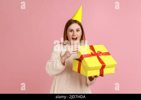 Porträt der aufgeregt erstaunt blonde Frau in Partei-Kegel Auspacken Geschenk-Box, mit angenehmer Überraschung, Blick auf die Kamera, trägt weißen Pullover. Innenaufnahme des Studios isoliert auf rosa Hintergrund. Stockfoto