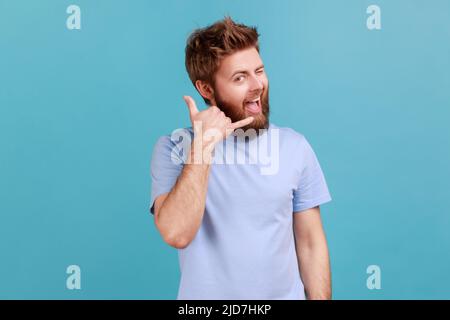 Rufen Sie mich an. Porträt eines positiv optimistischen bärtigen Mannes, der die Kamera anlächelt und mit einem Augenzwinkern zuwinkelt, der mich mit den Fingern zum Flirten bewegen lässt. Innenaufnahme des Studios isoliert auf blauem Hintergrund. Stockfoto