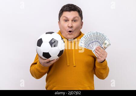 Überrascht Mann mittleren Alters zeigt Fußball und Spaß von hundert Dollar-Scheine, gewinnen viel Geld Wetten für Sport, tragen städtischen Stil Hoodie. Innenaufnahme des Studios isoliert auf weißem Hintergrund. Stockfoto