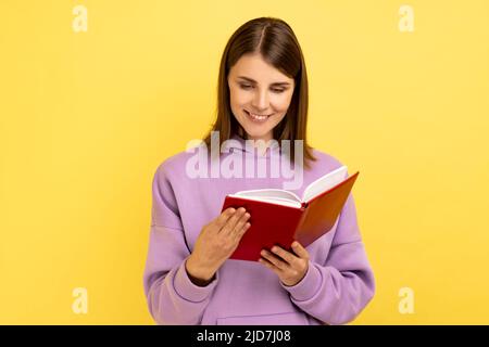 Portrait einer attraktiven lächelnden, konzentrierten Frau, die ein Interessantes Buch liest, sehr aufmerksam mit einer interessanten Handlung ist und einen violetten Hoodie trägt. Innenaufnahme des Studios isoliert auf gelbem Hintergrund. Stockfoto