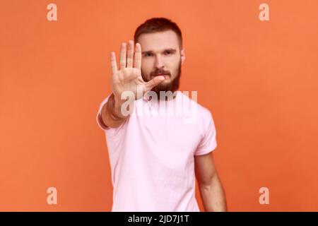 Ernst besorgter Mann macht Stop-Geste mit Handfläche, Konfliktverbotswarnung vor Gefahr, hört auf, Mobbing zu machen, trägt pinkes T-Shirt. Innenaufnahme des Studios isoliert auf orangefarbenem Hintergrund. Stockfoto
