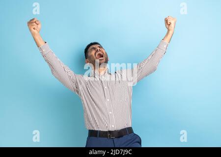 Porträt eines überglücklich aussehenden Geschäftsmannes, der mit erhobenen Fäusten und Schreien eine gewinnbringende Geste zum Ausdruck bringt, den Sieg feiert und ein gestreiftes Hemd trägt. Innenaufnahme des Studios isoliert auf blauem Hintergrund. Stockfoto