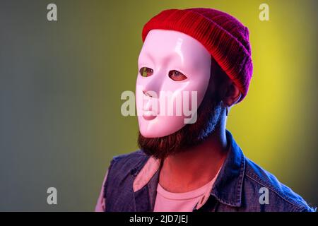 Seitenansicht des anonymen Hipster-Mannes in weißer Maske im Gesicht, versteckt seine Identität, spioniert Undercover, Korruption, trägt Hut und Jeansweste. Innenaufnahmen im Studio, isoliert auf farbigem Neonlicht-Hintergrund. Stockfoto