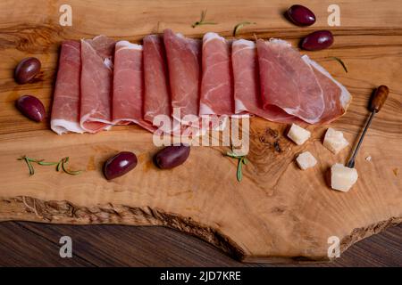 Draufsicht auf Scheiben italienischen Parmaschinkens, die auf einem rustikalen Holzschneidebrett mit Oliven, Parmesan-Flocken und Rosmarinblättern platziert sind Stockfoto