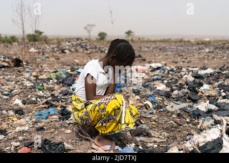 Erschöpftes kleines afrikanisches Mädchen kniet in einer Müllkippe, wo sie gezwungen wird, nach wiederverwendbarem Material zu suchen, Kinderarbeit und Ausbeutung