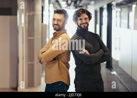 Portrait von glücklichen, gutaussehenden Unternehmern, die mit gekreuzten Armen posieren und in der Firmenhalle lächeln. Geschäftspartnerschaft Stockfoto