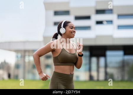 Sportliche junge schwarze Frau, die in Kopfhörern Musik hört, während sie auf der Straße joggt Stockfoto