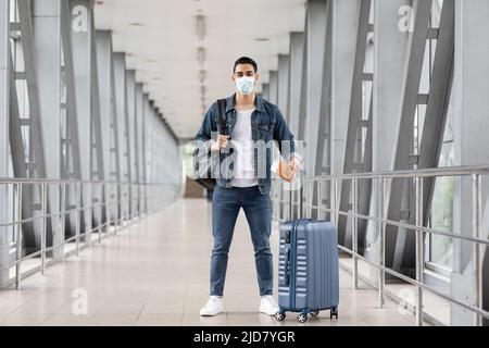 Junger Arabischer Mann Mit Medizinischer Gesichtsmaske, Der Mit Gepäck Im Flughafen Steht Stockfoto