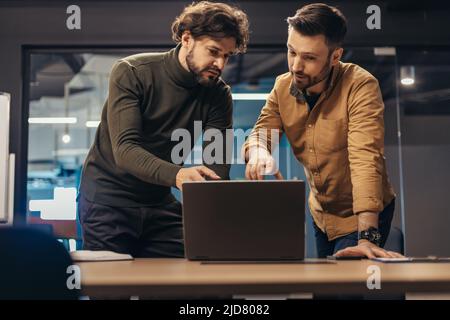 Portrait von zwei fokussierten männlichen Fachleuten, die gemeinsam an einem Startup-Projekt arbeiten, Ideen diskutieren und im Büro einen Laptop verwenden Stockfoto