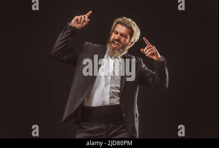 Fröhlicher, junger Geschäftsmann in schwarzem Anzug, der Musik mit Kopfhörern hört und auf grauem Hintergrund tanzt. Stockfoto