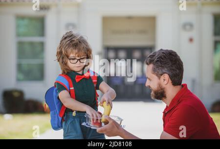 Vater unterstützt und motiviert Sohn. Kind geht zur Grundschule. Kleiner Schuljunge, der im Freien leckeres Mittagessen isst. Lehrertag. Stockfoto