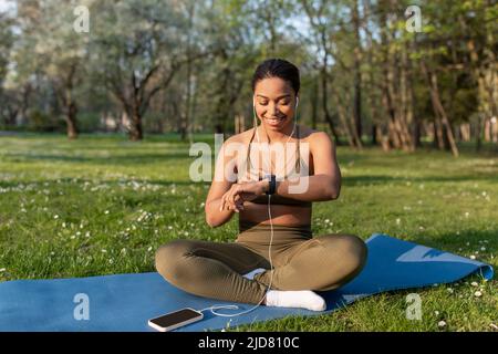 Junge schwarze Frau, die Mobiltelefon benutzt, Musik hört, Fitness Tracker während des Yoga-Trainings im Park überprüft Stockfoto