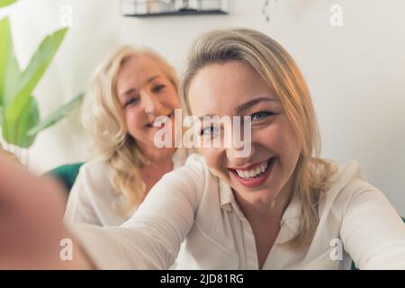 Blonde, europäische Frau mittleren Alters in einem weißen Hemd, die ein Selfie mit ihrer ähnlich aussehenden Mutter, die im Hintergrund steht, gemacht hat. Innenaufnahme. Hochwertige Fotos Stockfoto
