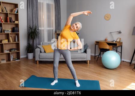 Glückliche ältere Frau, die zu Hause trainiert, fröhliche ältere Dame in Sportkleidung, die auf einer Yogamatte trainiert Stockfoto