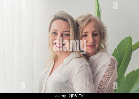 Schöne kaukasische blauäugige blonde Frauen aus dem Jahrtausend, die neben einem Fenster in einer hellen Wohnung stehen, die sich umarmt. Hochwertige Fotos Stockfoto