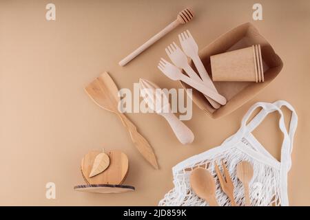 Umweltfreundliche Küchenutensilien, Einweggeschirr aus Papier und Netzbeutel. Holzbesteck und Einkaufstasche auf beigefarbenem Hintergrund. Nachhaltiger Lebensstil. Stockfoto