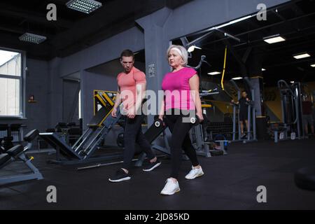 Ganzkörperaufnahme einer älteren Frau, die mit einem persönlichen Trainer gewichtete Ausfallschritte macht Stockfoto